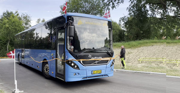 En blå bus med flag kører mellem to flagstænger, hvor der er udspændt en orange snor. På bussens display står der "Indvielsesfest"